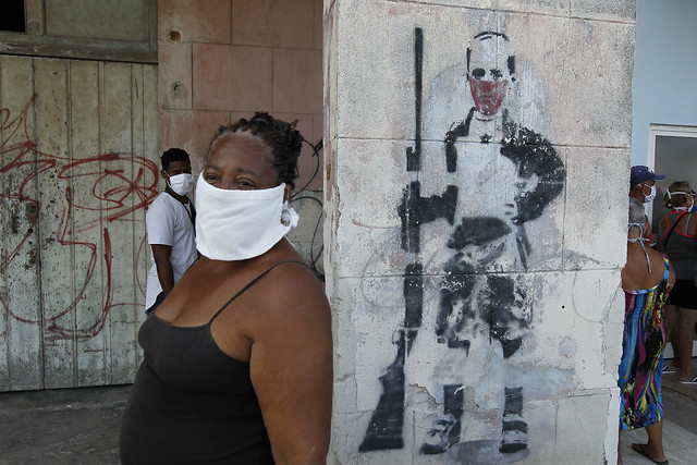 Una mujer negra, protegida con mascarilla para protegerse del contagio del coronavirus, espera para adquirir alimentos en el exterior de una tienda decorada con un grafiti callejero, en el municipio Playa, en La Habana, Cuba. Foto: Jorge Luis Baños/IPS